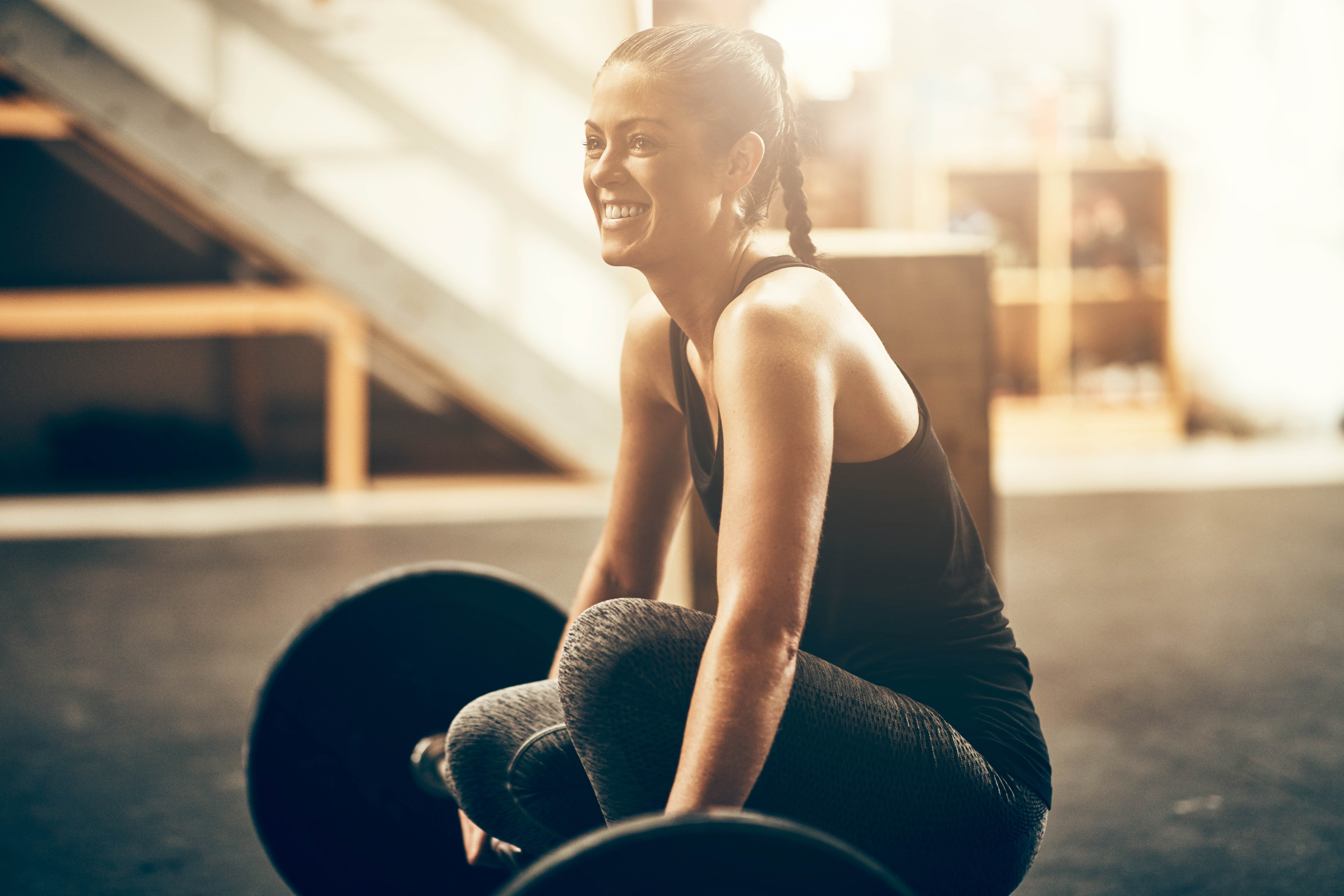 Young woman smiling as she kneels down next to her barbell in a gym Young woman smiling as she kneels down next to her barbell in a gym
